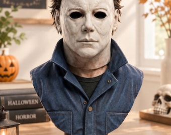 May include: A bust of a character wearing a white, weathered mask with dark eye sockets and brown hair. The figure is dressed in a blue collared jacket over a black shirt, mounted on a decorative brown base with an empty rectangular nameplate.