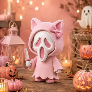May include: A stylized figurine featuring a white ghost-like mask with elongated eye and mouth openings, wearing a light pink hooded cloak with cat ears and a small bow. The figure holds a small white knife in its right hand.