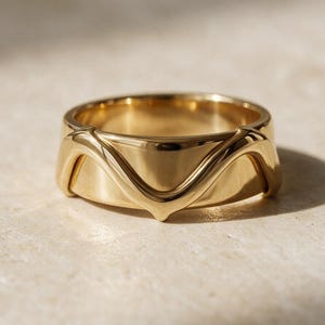May include: A polished gold band ring featuring a smooth, continuous surface with a raised, wave-like V-shaped detail that wraps around the exterior. The metal has a reflective, high-shine finish that catches the light, creating distinct highlights.