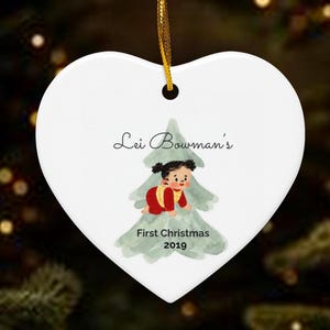 K&ouml;nnte beinhalten: Ein wei&szlig;er, herzf&ouml;rmiger Keramikschmuck mit einer goldenen Aufh&auml;ngeschnur. Er zeigt eine Aquarellillustration eines Babys in roter Kleidung vor einem gr&uuml;nen Baum mit dem aufgedruckten Text Lei Bowman's First Christmas 2019.