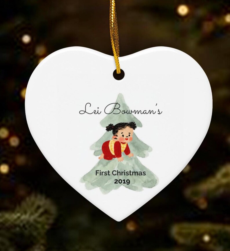 K&ouml;nnte beinhalten: Ein wei&szlig;er, herzf&ouml;rmiger Keramikschmuck mit einer goldenen Aufh&auml;ngeschnur. Er zeigt eine Aquarellillustration eines Babys in roter Kleidung vor einem gr&uuml;nen Baum mit dem aufgedruckten Text Lei Bowman's First Christmas 2019.