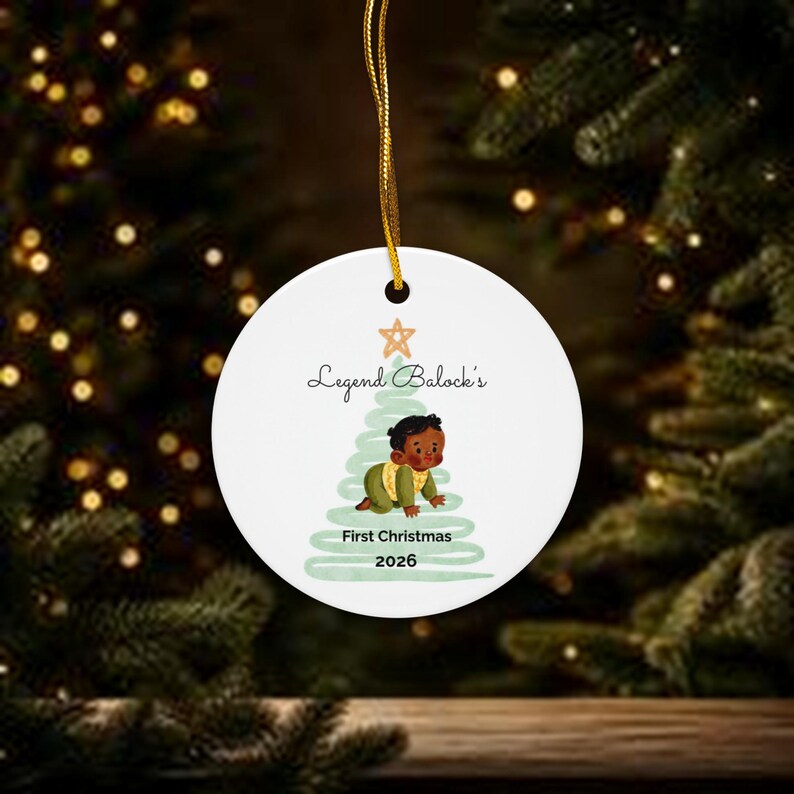 K&ouml;nnte beinhalten: Ein wei&szlig;er, runder Keramik-Weihnachtsbaumschmuck an einer goldenen Schnur. Das Motiv zeigt einen hellgr&uuml;nen Aquarell-Weihnachtsbaum, einen Stern und ein krabbelndes Baby. Der Text lautet: Legend Balock's First Christmas 2026.
