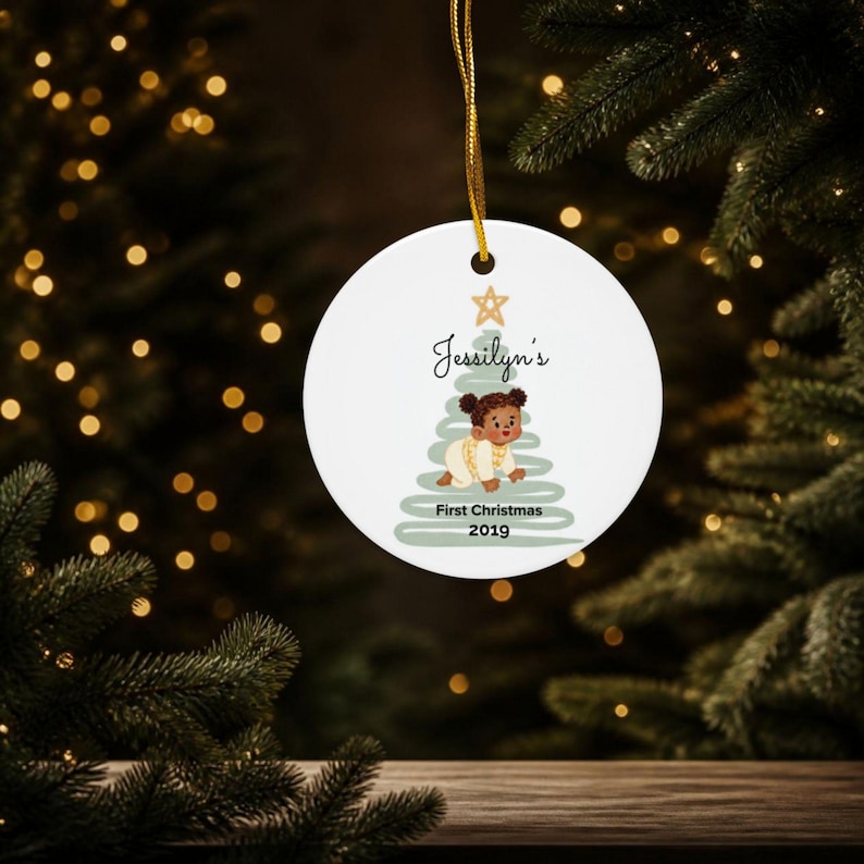 May include: A round white ceramic ornament hanging by a gold string. The design features a stylized sage green Christmas tree with a gold star, a crawling baby illustration, and the text Jessilyn's First Christmas 2019 in black script.