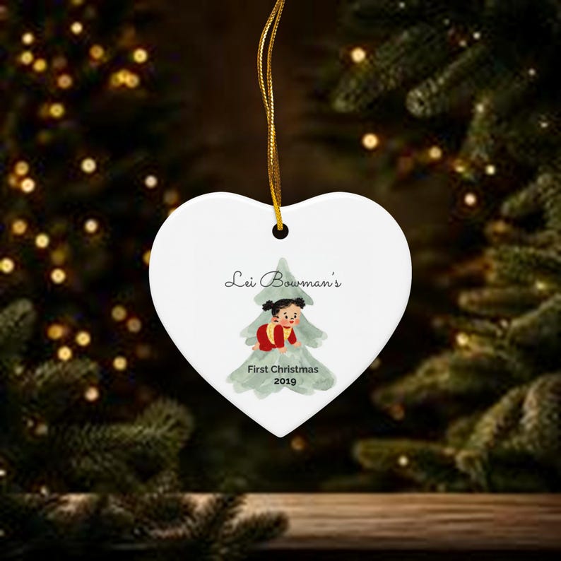 K&ouml;nnte beinhalten: Ein wei&szlig;er herzf&ouml;rmiger Keramikschmuck, der an einer goldenen Schnur h&auml;ngt. In der Mitte befindet sich eine gr&uuml;ne Aquarell-Weihnachtsbaumgrafik mit einem kleinen Kind in roter Kleidung und dem Text Lei Bowman's First Christmas 2019.