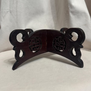 May include: A dark brown wooden display stand featuring an X-shaped base with four curved legs. The center of each side displays a circular cutout with an intricate, symmetrical geometric pattern carved into the wood surface.