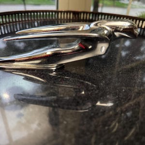May include: A polished chrome hood ornament featuring a stylized, aerodynamic figure in a horizontal, outstretched pose. The metallic surface is highly reflective, showing smooth, sweeping lines that taper toward the front of the object.