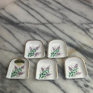 May include: A set of five small white ceramic dishes with a gold-trimmed edge and a hand-painted floral motif featuring green leaves and purple blossoms. One dish includes a gold sticker with the Japanese text 清水焼, indicating Kiyomizu-yaki.