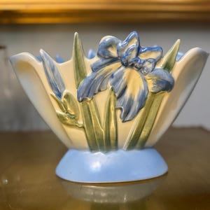 May include: A cream-colored ceramic bowl featuring a raised, three-dimensional relief of blue iris flowers and green leaves. The bowl has a scalloped rim and sits on a solid light blue circular base, with a smooth, glossy finish throughout the piece.