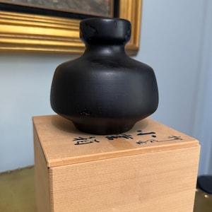May include: A matte black ceramic vase with a wide, rounded body and a narrow neck, resting on a light-colored wooden box. The top of the wooden box features black ink calligraphy characters written in a vertical orientation across the surface.