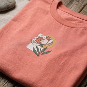 May include: A coral-colored t-shirt featuring a graphic print of a white and pink peony flower with green leaves, positioned over a light rectangular background and a yellow circle. The shirt has a crew neckline and a soft, textured fabric finish.
