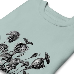 May include: A light sage green t-shirt featuring a detailed black and white botanical illustration of various pitcher plants. The graphic displays multiple carnivorous plants with distinct cup-shaped structures and spiky edges arranged in a cluster.