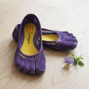 Pode incluir: Um par de sapatos Vibram FiveFingers roxos com cabedal em malha, compartimentos individuais para os dedos e palmilha interna amarelo-brilhante. A marca Vibram está impressa em amarelo na lateral e na palmilha deste calçado flexível.