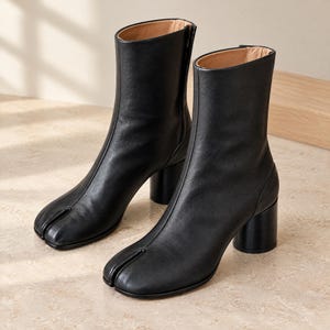 May include: A pair of black leather ankle boots featuring a distinct split-toe design, a vertical seam running up the front, and a sturdy cylindrical block heel. The interior of the boots is lined with a light tan material, and the exterior has a smooth finish.