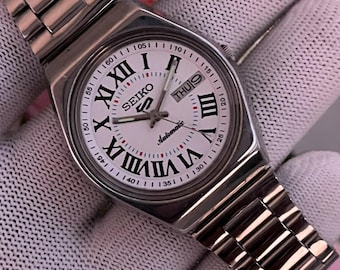 May include: A silver-tone Seiko 5 automatic wristwatch with a white dial, black Roman numeral hour markers, and a stainless steel link bracelet. The watch face features the Seiko logo, the number 5 emblem, and a day-date window at the 3 o'clock position.