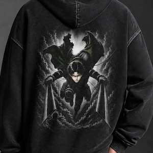 Puede incluir: Una sudadera con capucha de color negro carbón con efecto lavado al ácido y un gran estampado gráfico en la espalda. El diseño muestra un personaje de anime con una capa verde oscuro, blandiendo dos hojas entre humo y efectos de luz.