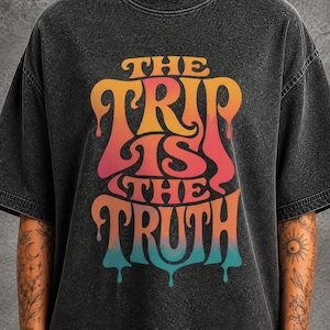 May include: A charcoal grey acid-wash t-shirt featuring a graphic print with the text "THE TRIP IS THE TRUTH" in a stylized, dripping font. The lettering transitions through colors of orange, red, and teal against the dark fabric background.