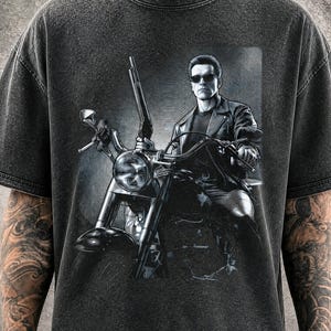 Könnte beinhalten: Ein anthrazitfarbenes T-Shirt in Acid-Wash-Optik mit einem Grafikdruck einer Figur in Lederjacke und Sonnenbrille auf einem Motorrad, die eine Schrotflinte hält. Das Design zeigt ein kreisförmiges radiales Linienmuster im Hintergrund.