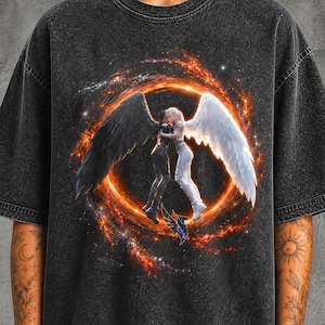 Może przedstawiać: Ciemnoszary t-shirt z efektem sprania, ozdobiony grafiką przedstawiającą dwie postacie obejmujące się wewnątrz płonącego pomarańczowego pierścienia. Jedna postać ma duże czarne skrzydła, a druga duże białe skrzydła. Poniżej znajduje się mały niebieski ptak.