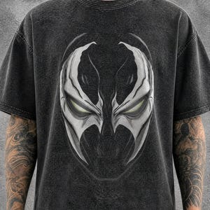 以下が含まれることがあります： チャコールブラックのアシッドウォッシュ加工Tシャツ。中央には、鋭く角張った特徴と輝く黄色い目を持つ、白とグレーの様式化されたマスクの大きなグラフィックが描かれています。クルーネック、半袖、ゆったりとしたボックスシルエットのデザインです。