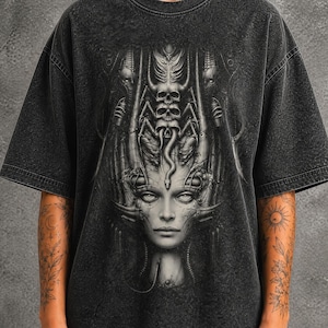 Könnte beinhalten: Ein anthrazitfarbenes T-Shirt in Acid-Wash-Optik mit einem detaillierten, einfarbigen Grafikdruck auf der Brust. Das Design zeigt ein zentrales, menschenähnliches Gesicht, umgeben von komplexen biomechanischen Strukturen, Totenköpfen und schlangenartigen Elementen.