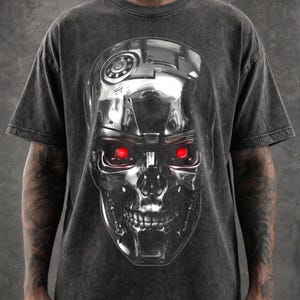 Könnte beinhalten: Ein anthrazitfarbenes T-Shirt in Acid-Wash-Optik mit einer großen, detaillierten Grafik eines metallischen Roboterschädels. Der Schädel hat eine polierte Chromoberfläche mit komplexen mechanischen Details und leuchtend roten Augen.