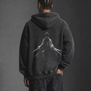 May include: A charcoal grey acid-wash hooded sweatshirt featuring a white graphic print on the back. The illustration depicts a figure viewed from behind, wearing a flowing cloak with detailed line work. The hoodie has long sleeves and ribbed cuffs.