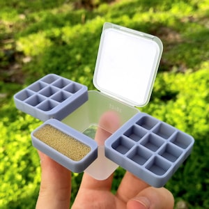 以下が含まれることがあります： ヒンジ付きの蓋と3つのコンパートメントを備えた透明なプラスチック製収納容器。2つのコンパートメントには9つの小さな正方形のグリッドがあり、3つ目のコンパートメントには質感のある明るいベージュ色のフォームインサートが入っています。