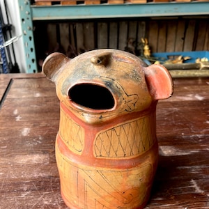 Puede incluir: Un recipiente cilíndrico de terracota con una boca ancha y abierta y dos asas que sobresalen en forma de orejas. La superficie está decorada con patrones geométricos negros dibujados a mano sobre un acabado de arcilla naranja y marrón, creando una forma animal.