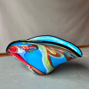 Puede incluir: Un cuenco de vidrio soplado a mano con una base de color azul turquesa vibrante y patrones de remolinos en rojo, verde y blanco. El cuenco tiene una forma asimétrica y curva con un borde negro liso y pulido que perfila la parte superior.