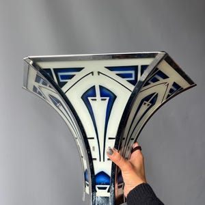 May include: A wall sconce light fixture featuring a wide, fan-shaped glass shade with a white background and bold, geometric blue patterns. The glass panels are framed by polished chrome metal edges that create a structured, angular appearance.