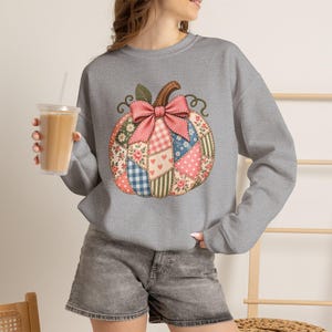 May include: A heather grey long-sleeve sweatshirt featuring a large central graphic of a pumpkin made of various patchwork patterns including floral, gingham, polka dot, and heart prints, topped with a prominent pink bow and green leaves.