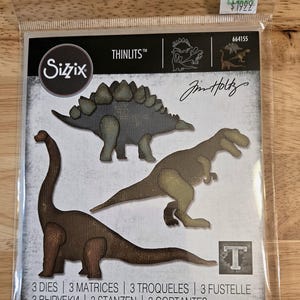May include: A Sizzix Thinlits die set by Tim Holtz in clear plastic packaging featuring three dinosaur shapes: a Stegosaurus, a Tyrannosaurus Rex, and a long-necked sauropod. The package includes the text Sizzix, Thinlits, and 3 Dies.