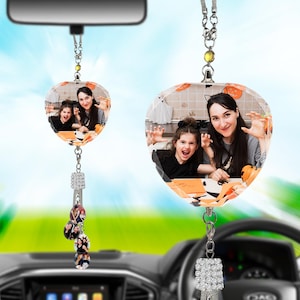 Op de afbeelding: Een hartvormige autospiegelhanger met een afgedrukte foto van een persoon en een kind met Halloween-decoraties. De hanger hangt aan een zilveren ketting met een met strass ingelegde bedel en vier kleinere ronde fotohangers.