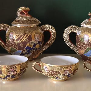 May include: A tea set featuring a teapot and a lidded sugar bowl with dragon-shaped handles and finials, three teacups, and several saucers. The pieces are decorated with intricate gold, brown, and blue patterns depicting figures and ornate motifs.