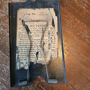 May include: A vintage black metal McCaskey account register clip with a spring-loaded wire mechanism. The center features a weathered, torn paper insert with printed text addressed to patrons, including the heading TO OUR PATRONS and Reg. No. 37.