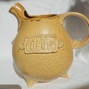 May include: A tan ceramic pitcher with a rounded body, a curved handle, and a spout. The front features a raised rectangular plaque with the embossed text JOSE CUERVO. The lower portion of the pitcher has a textured, cross-hatched pattern.