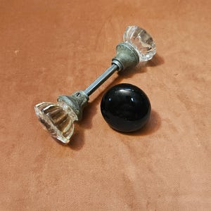 May include: A clear, faceted glass door knob set connected by a metal spindle and a single, smooth, round, solid black door knob. The glass knobs feature a multi-sided, reflective design with weathered metal bases attached to the central rod.