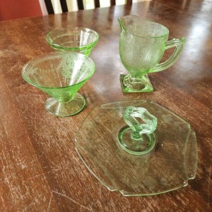 Puede incluir: Un juego de cuatro piezas de vidrio verde claro que incluye una jarra de crema con asa, dos cuencos con pie y patrones grabados, y una bandeja de servicio plana con un asa central en forma de bucle. Cada pieza tiene un acabado de vidrio verde.