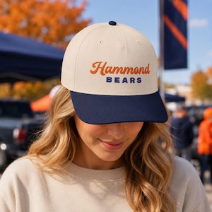 May include: A cream-colored baseball cap with a dark navy blue brim and a dark navy blue button on top. The front of the cap features the text Hammond in orange cursive script above the word BEARS in dark navy blue block capital letters.