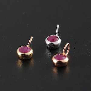May include: Three small circular pendants, each featuring a faceted round pink gemstone set in metal. One pendant is yellow gold, one is silver-toned, and one is rose gold. Each piece has a small attached jump ring and is stamped with the text AU750.