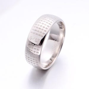 May include: A polished silver-toned metal band ring featuring a wide, flat surface engraved with intricate rows of small, dark Chinese characters. The ring has a smooth, reflective finish and a rounded interior profile, showcasing a continuous script.