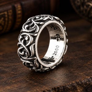 Puede incluir: Un anillo de metal plateado con un intrincado patrón de volutas en relieve en la banda exterior. El interior de la banda es liso y está claramente grabado con el texto 2000 junto con pequeños símbolos en forma de cruz en el borde interior.