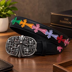 May include: A black textured leather belt featuring a series of colourful embroidered cross patches in green, orange, pink, purple, and red. The belt includes a large, oval-shaped silver-toned metal buckle with an intricate embossed cross design.