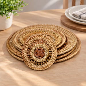 May include: A stack of round, woven rattan coasters featuring a light tan natural finish and a darker brown decorative knot pattern at the center of each piece. The coasters are circular with a detailed, intricate braided texture throughout.