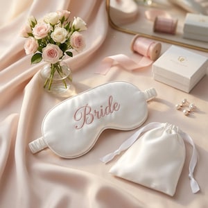Peut inclure: Un masque de sommeil en satin blanc avec le mot Bride brodé en écriture rose clair, accompagné d'une pochette à cordon en satin blanc, d'une petite boîte cadeau blanche, d'une paire de boucles d'oreilles en perles et d'un vase en verre.