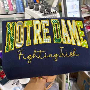 May include: A navy blue sweatshirt featuring the text NOTRE DAME in large, block-style letters with various green and white patterned fabric appliques outlined in yellow thread, and the text Fighting Irish embroidered in yellow cursive below.