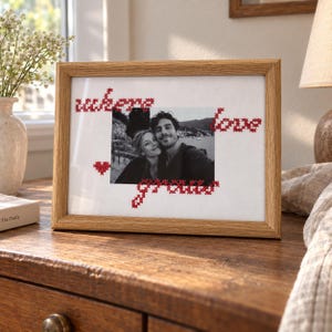 Puede incluir: Un marco de fotos rectangular de madera clara que contiene una fotografía en blanco y negro de una pareja. El paspartú blanco presenta texto bordado en punto de cruz rojo que dice where love grows, junto a un pequeño corazón.