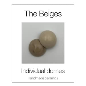 May include: Two smooth, semi-spherical ceramic domes in varying shades of beige. The text "The Beiges", "Individual domes", and "Handmade ceramics" is printed in black on the white background surrounding the two ceramic objects.