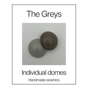 May include: Two circular ceramic domes labelled The Greys Individual domes Handmade ceramics. One dome is a smooth, glossy light grey, while the other is a textured, matte dark charcoal grey. Both objects are round, convex, and solid in appearance.