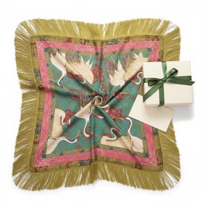May include: A square scarf with a teal background featuring white crane birds and red floral patterns, bordered by a pink frame and long olive green fringe. A cream-colored gift box with a dark green ribbon and a blank card are placed on top.